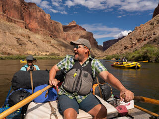 A man with a beard is smiling as he rows a raft on the Colorado River in the Grand Canyon.