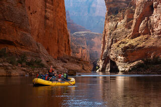 A yellow raft floats the Colorado River surrounded by tall colorful cliffs.