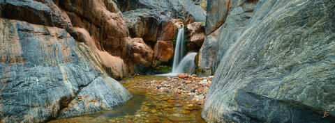 Clear Creek Falls in the Grand Canyon is a waterfall surrounded by gray, blue, and peach colored cliffs.