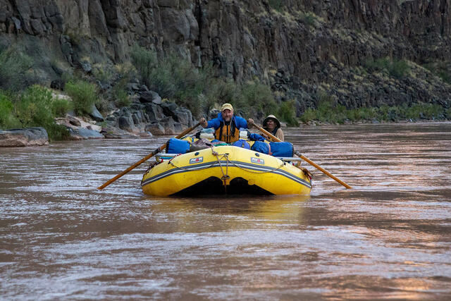 Person rowing a yellow raft on calm water in a canyon.