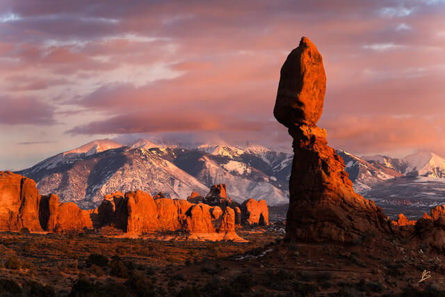 Golden light on Balanced Rock in Arches National Park with snow-covered mountains in the distance and colorful clouds at sunset.