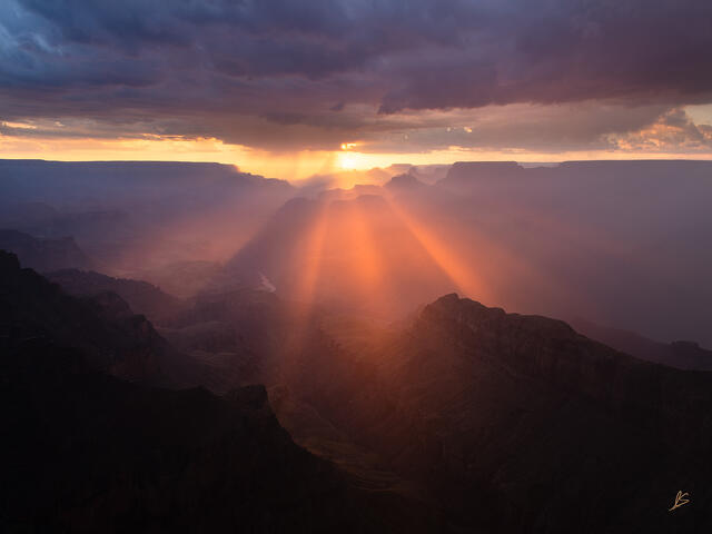 Colorful beams of sunlight in the Grand Canyon at sunset with dark storm clouds overhead.