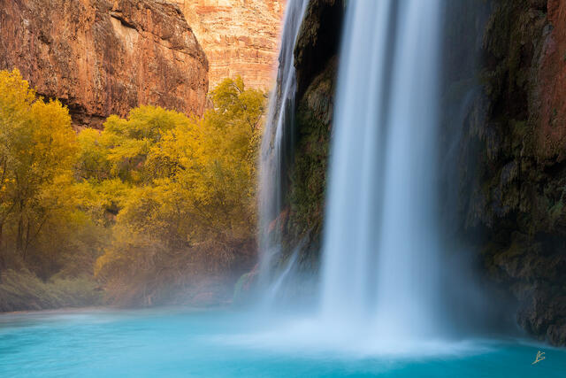 A waterfall cascades into a pool of turquoise water surrounded by trees with yellow leaves and redrock cliffs.