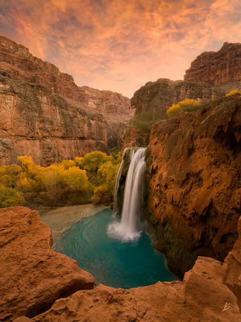 Red rock cliffs and fall color surround Havasu Falls at sunrise with a beautiful sky full of colorful clouds.