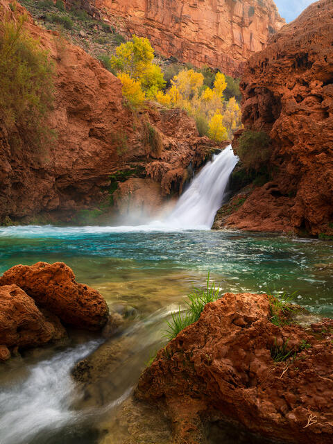 Orange and red cliffs and trees with yellow leaves surround a beautiful waterfall cascading into a pool of blue-green water.