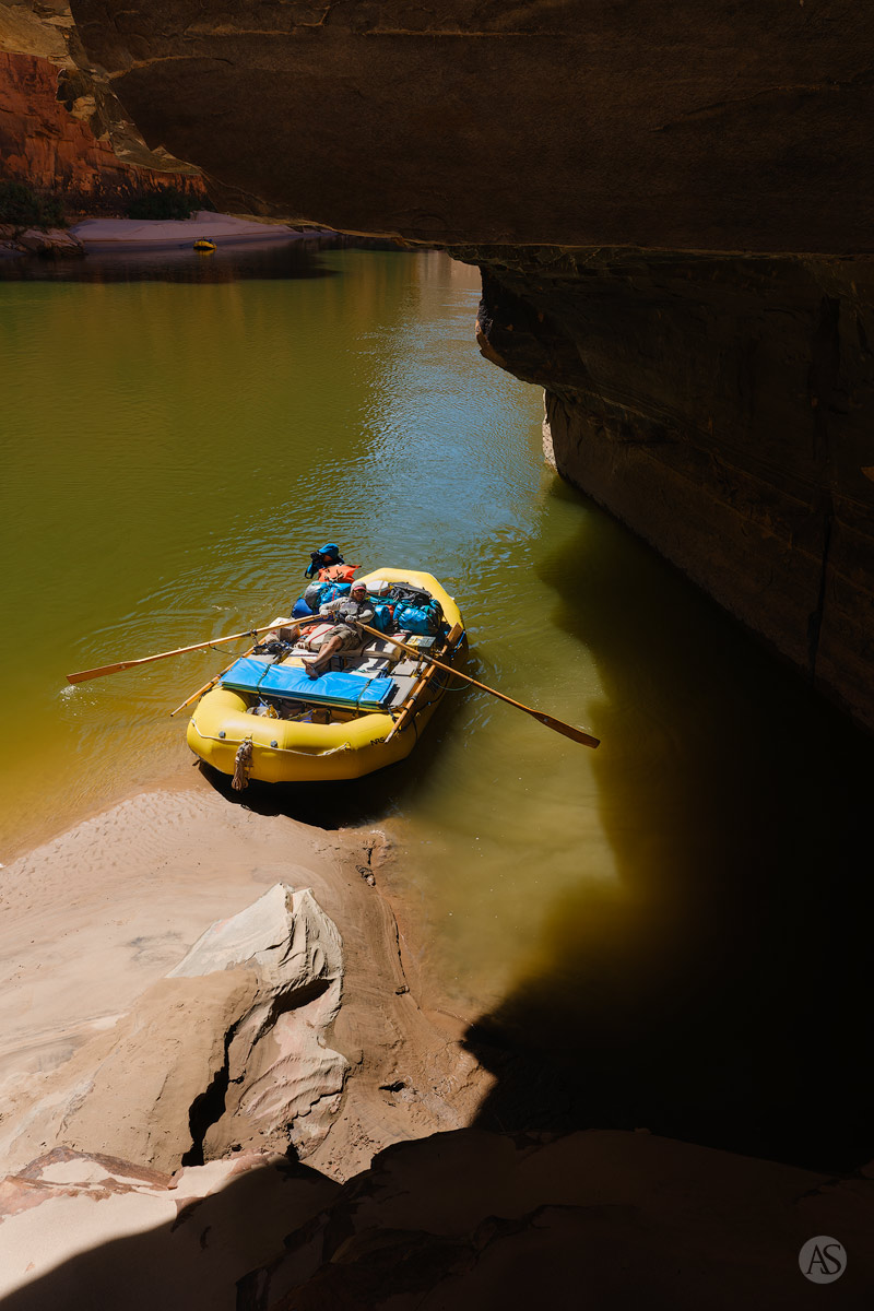 A yellow raft floats on the Colorado River below an overhanging alcove in the Grand Canyon.