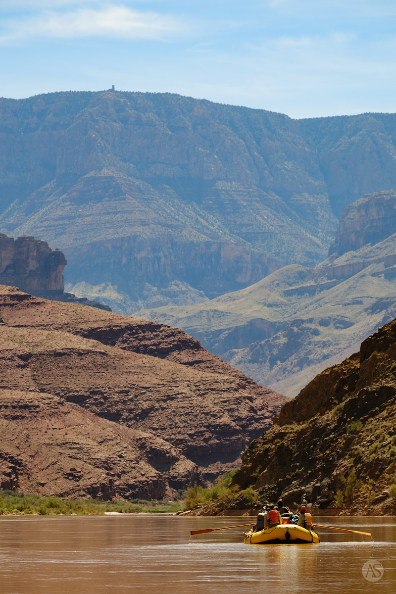 A yellow raft floats along the Colorado River with the Desert View Watchtower in the distance at the top of the Grand Canyon.