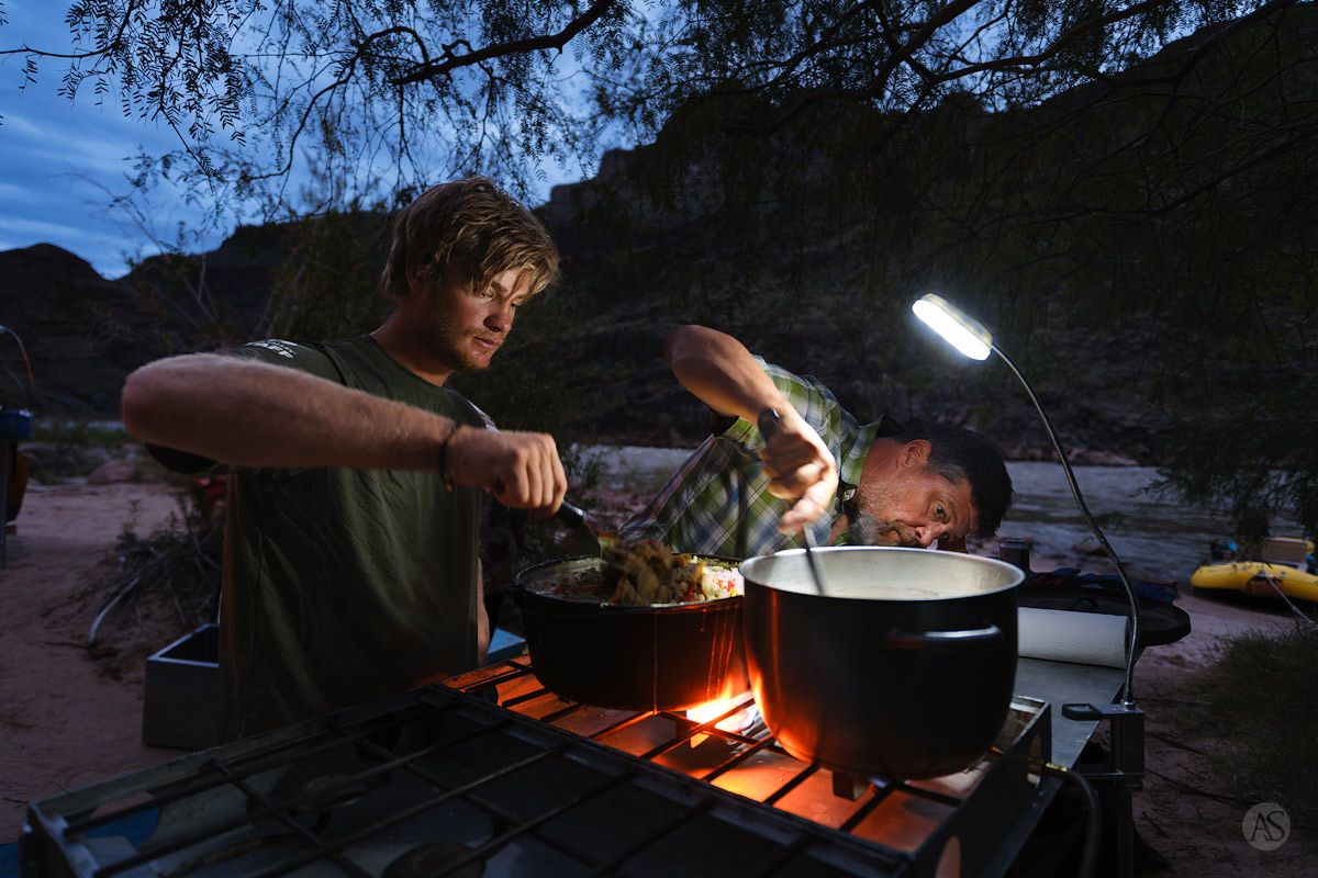 Two river guides preparing dinner in a pot over a flame at night while camping in the Grand Canyon.