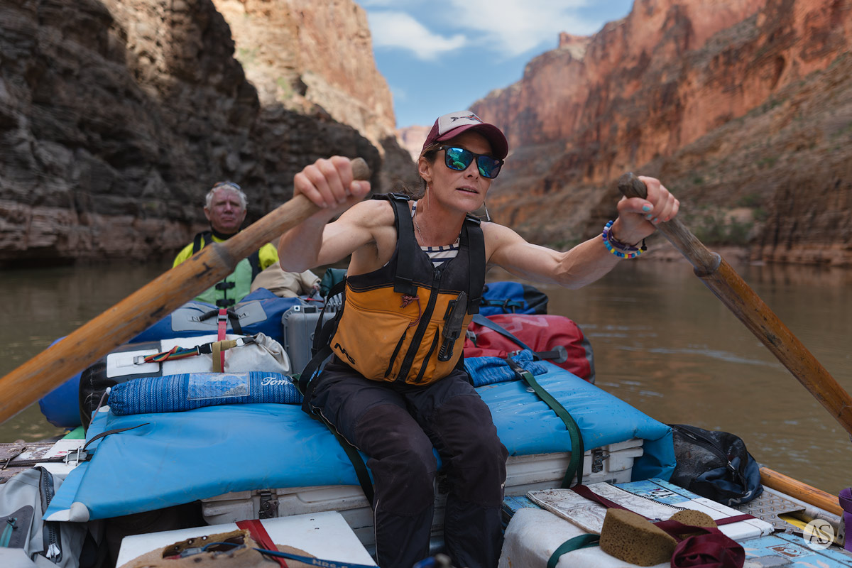 A female river guide with her hands on the oars stares downstream, studying the current of the Colorado River in the Grand Canyon as a passenger looks on.