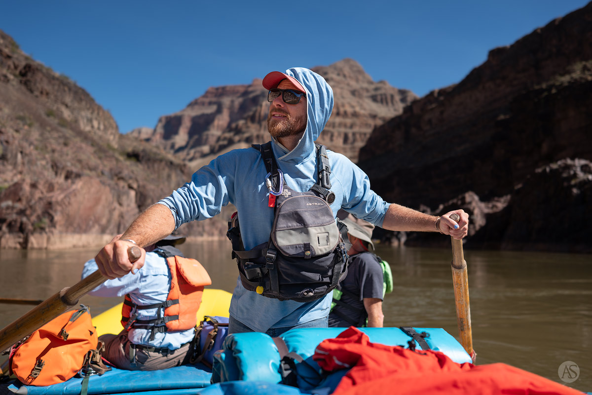 A river guide in a blue hoodie with his hands on the oars enjoys the view of the Grand Canyon as he guides his raft downstream on the Colorado River.